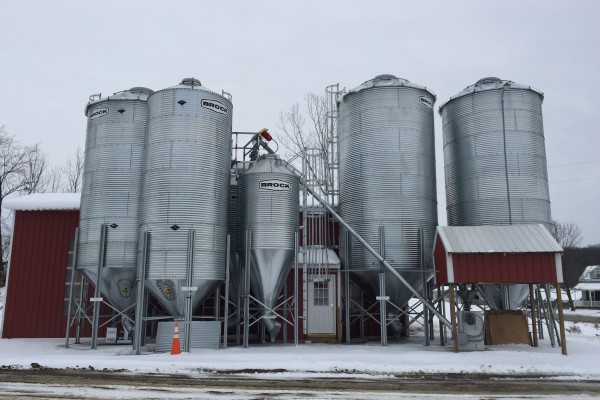 slim Brock grain bins with red farm buildings in front and behind them on a snowy day
