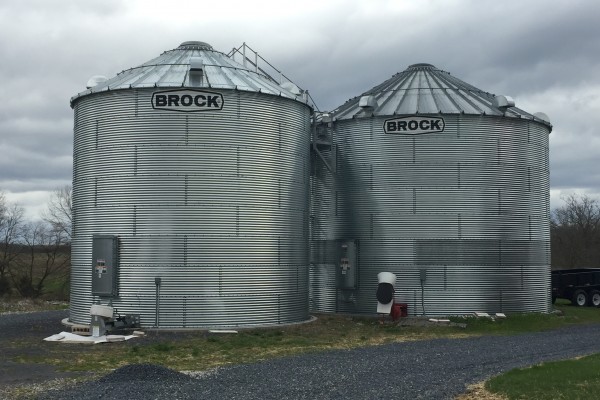two Brock grain bins next to each other with a gravel road passing in front of them