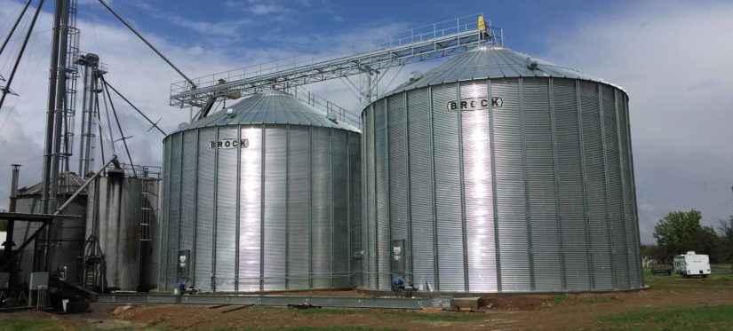 two Brock grain bins next to a farm system in a rural area on a beautiful day